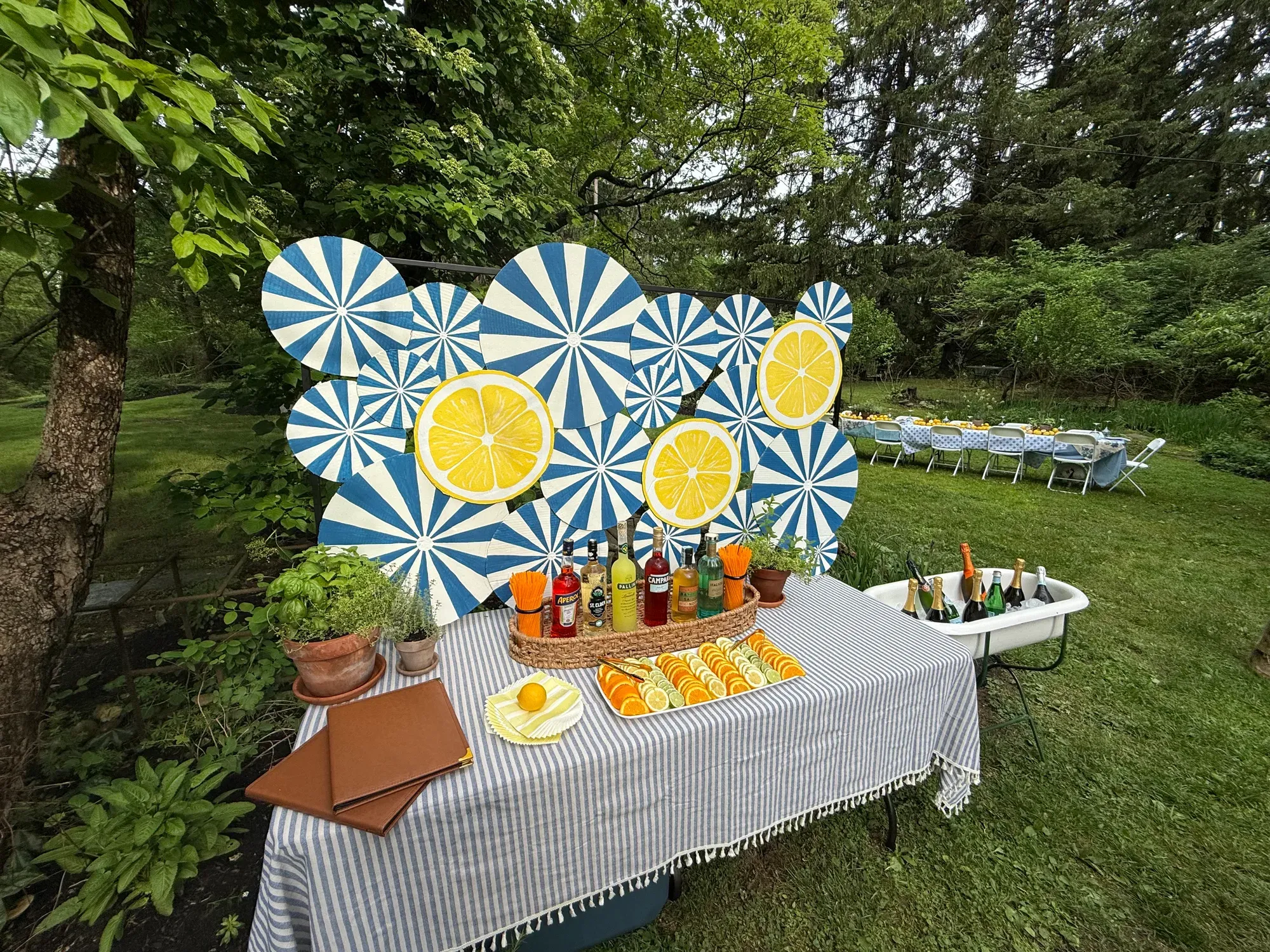An aperitivo bar in a garden: bottles, sliced citrus and potted herbs on a striped cloth, in front of a blue-and-white backdrop with painted lemons, and a long table set behind.