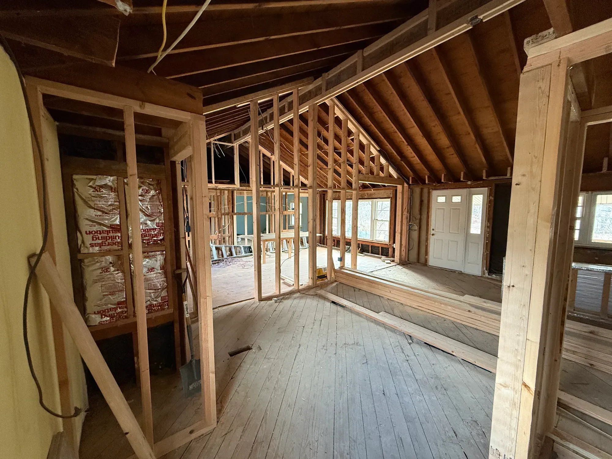 A guest house mid-rehab: open stud walls, exposed rafters and insulation, with daylight from a row of windows.